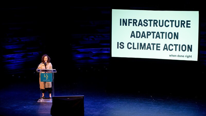 Anya Domlesky behind a podium on a stage with a large screen next turn her show a white slide with bold black words "INFRASTRUCTURE ADAPTATION IS CLIMATE ACTION when done right"
