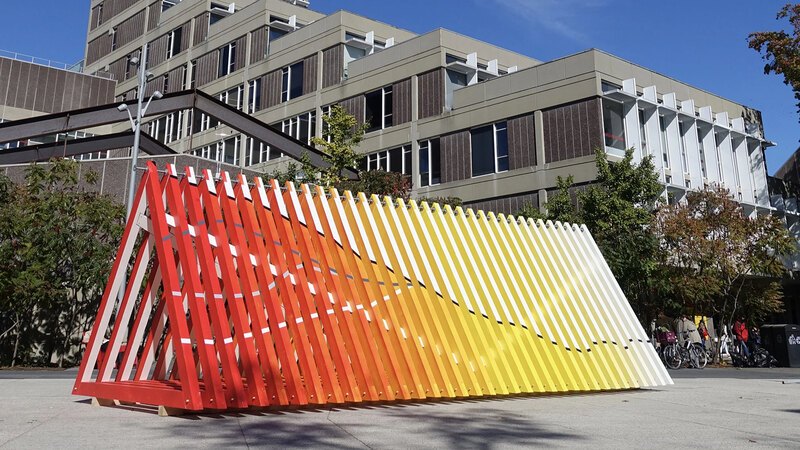 A colorful triangular tunnel of wood planks serves as an art installation in a plaza